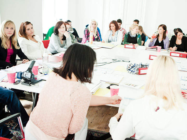 Eine Gruppe von Menschen sitzt in einem hellen Raum um einen Konferenztisch. Sie sind in eine Besprechung vertieft. Auf dem Tisch liegen Papiere, Ordner und rosa Becher. Die Teilnehmer wirken aufmerksam und konzentriert.