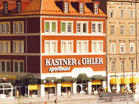 In einem historischen Gebäude befindet sich Kastner & Öhler, ein Kaufhaus mit roter Fassade und weißen Akzenten. Große Fenster haben grüne Fensterläden. Fußgänger gehen vorbei, und auf Straßenebene befinden sich Geschäfte mit gelben Markisen.
