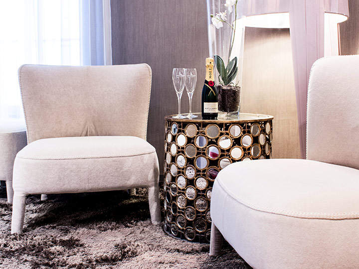 Zwei elegante beige Stühle stehen um einen kleinen runden Tisch mit einem dekorativen Metallfuß. Auf dem Tisch stehen eine Flasche Champagner, zwei Gläser und eine weiße Orchidee in einem Topf. Der Tisch steht auf einem weichen grauen Teppich mit sanfter Beleuchtung im Hintergrund.