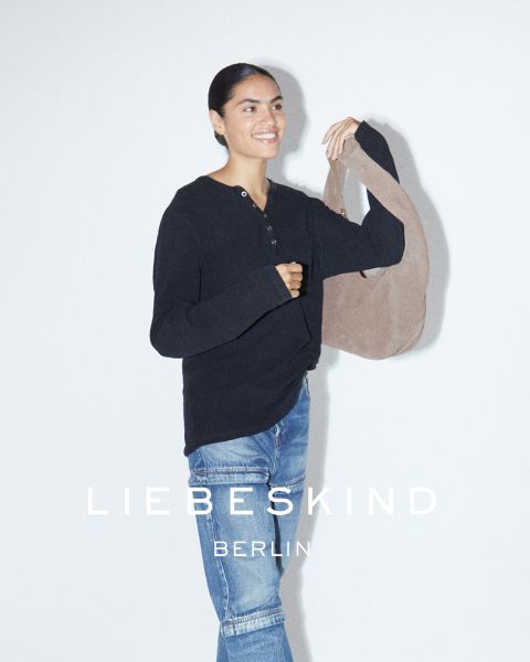 Eine lächelnde Frau in einem schwarzen langärmeligen Hemd und Jeans trägt eine hellbraune Tasche über der Schulter. Der Text "Liebeskind Berlin" erscheint am unteren Rand des Bildes.