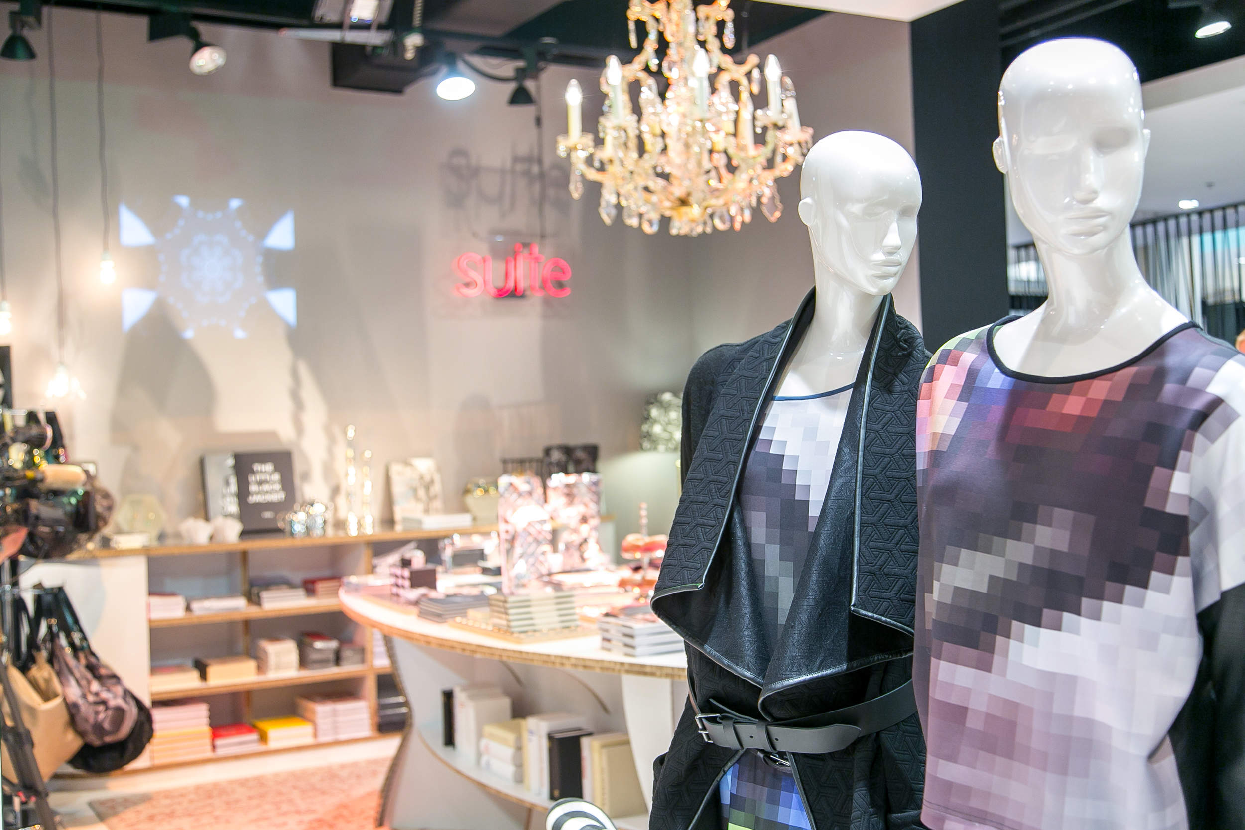 Schaufensterpuppen in Outfits mit Pixelmustern stehen in einem stilvollen Laden, der Bücher, Taschen und Dekorationen ausstellt. Darüber hängt ein Kronleuchter, und an der Wand ist ein Neonschild mit der Aufschrift „Suite“ zu sehen.
