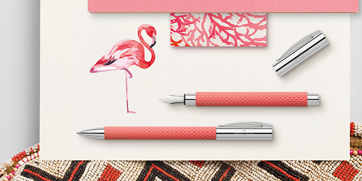 Rosa und silberne Stifte liegen auf einer weißen Oberfläche neben einer Zeichnung eines Flamingos und einer Karte mit Korallenmuster. Unten ist teilweise ein Perlengegenstand mit geometrischen Mustern sichtbar.