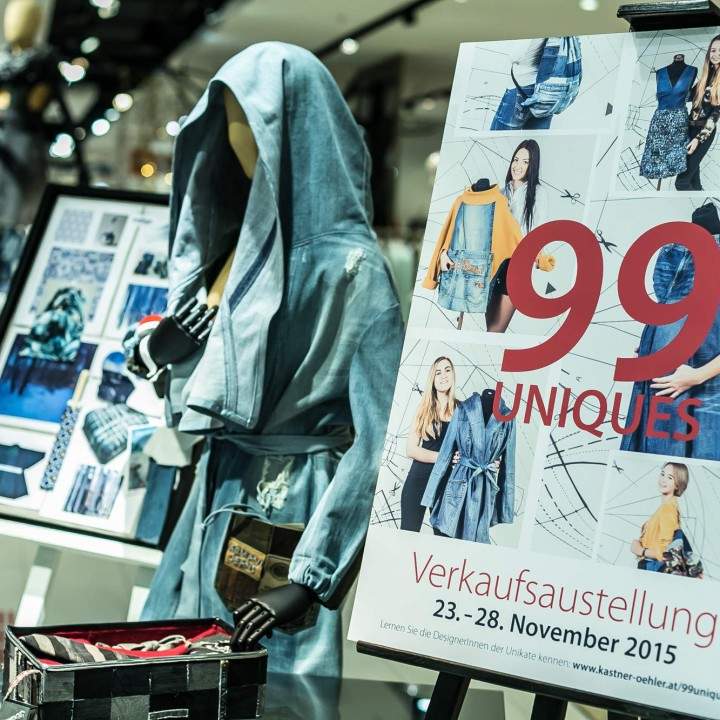 Auf einem Schaufenster sind eine Jeansjacke und ein Werbeschild für „99 Uniques“ mit Bildern verschiedener Outfits zu sehen. Die Veranstaltung findet vom 23. bis 28. November 2015 statt. Das Schaufenster befindet sich in einem hell erleuchteten Einzelhandelsumfeld.