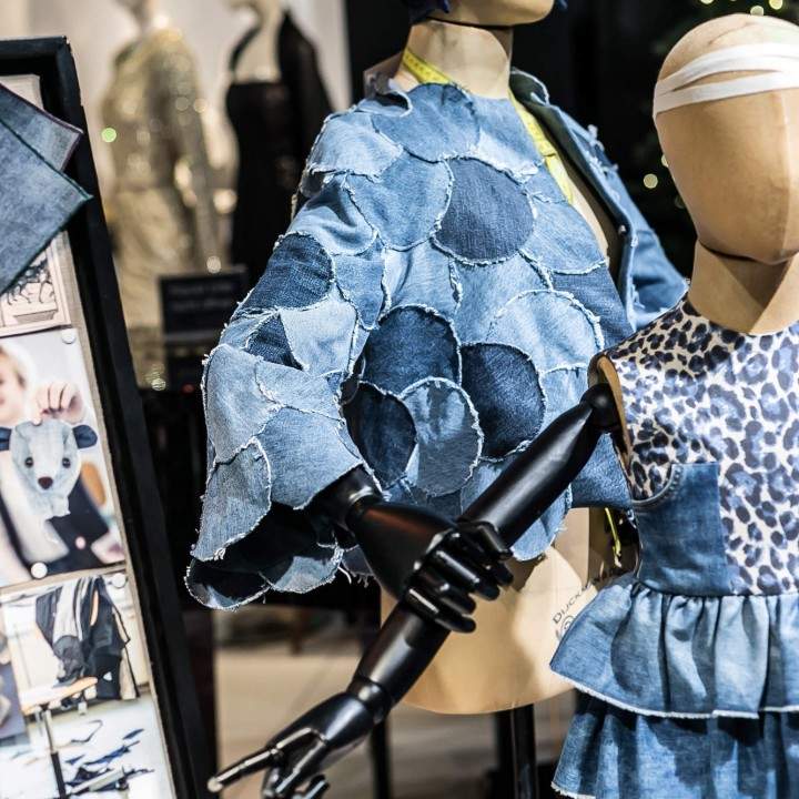 Ausstellung von Schaufensterpuppen in modischen Jeans-Outfits, darunter eine strukturierte Jacke und ein Lagenkleid mit Animalprint. Daneben zeigt eine Tafel Stoffmuster und Designfotos. Eine moderne und kreative Designausstellung.