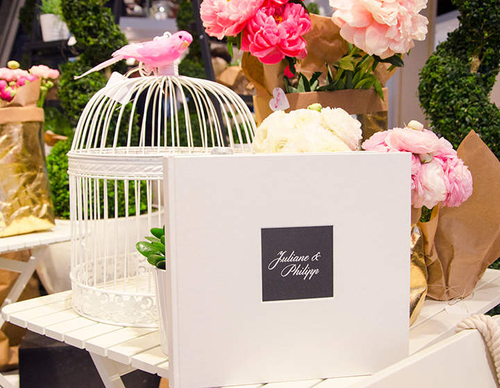 Ein dekorierter Tisch mit rosa und weißen Blumen, ein weißer Vogelkäfig, der mit einem kleinen rosa Vogel geschmückt ist, und ein weißes Buch mit einem schwarzen Etikett mit der Aufschrift „Juliane & Philipp“.