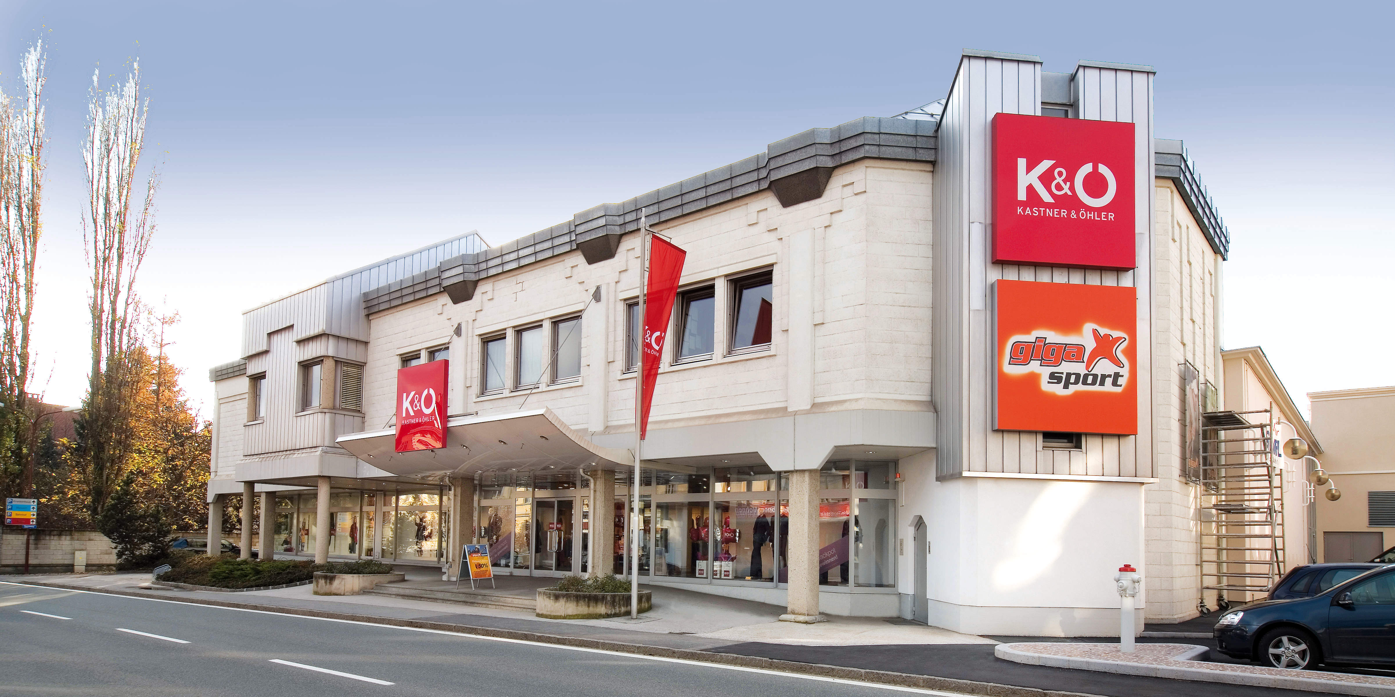 Ein modernes zweistöckiges Gebäude mit großen Fenstern. Es verfügt über rote Schilder für „Kastner & Öhler“ und „giga X sport“. Die Fassade enthält dekorative Säulen und das Gebäude liegt neben einer breiten Straße.