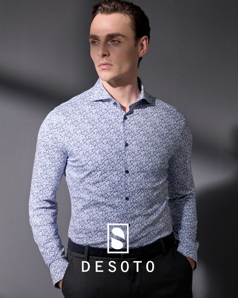 Ein Mann mit einem langärmeligen, gemusterten Hemd und einer dunklen Hose steht vor einem grauen Hintergrund. Er blickt mit den Händen in den Taschen zur Seite. Der Schriftzug "DESOTO" und das DESOTO-Logo sind am unteren Rand zu sehen.