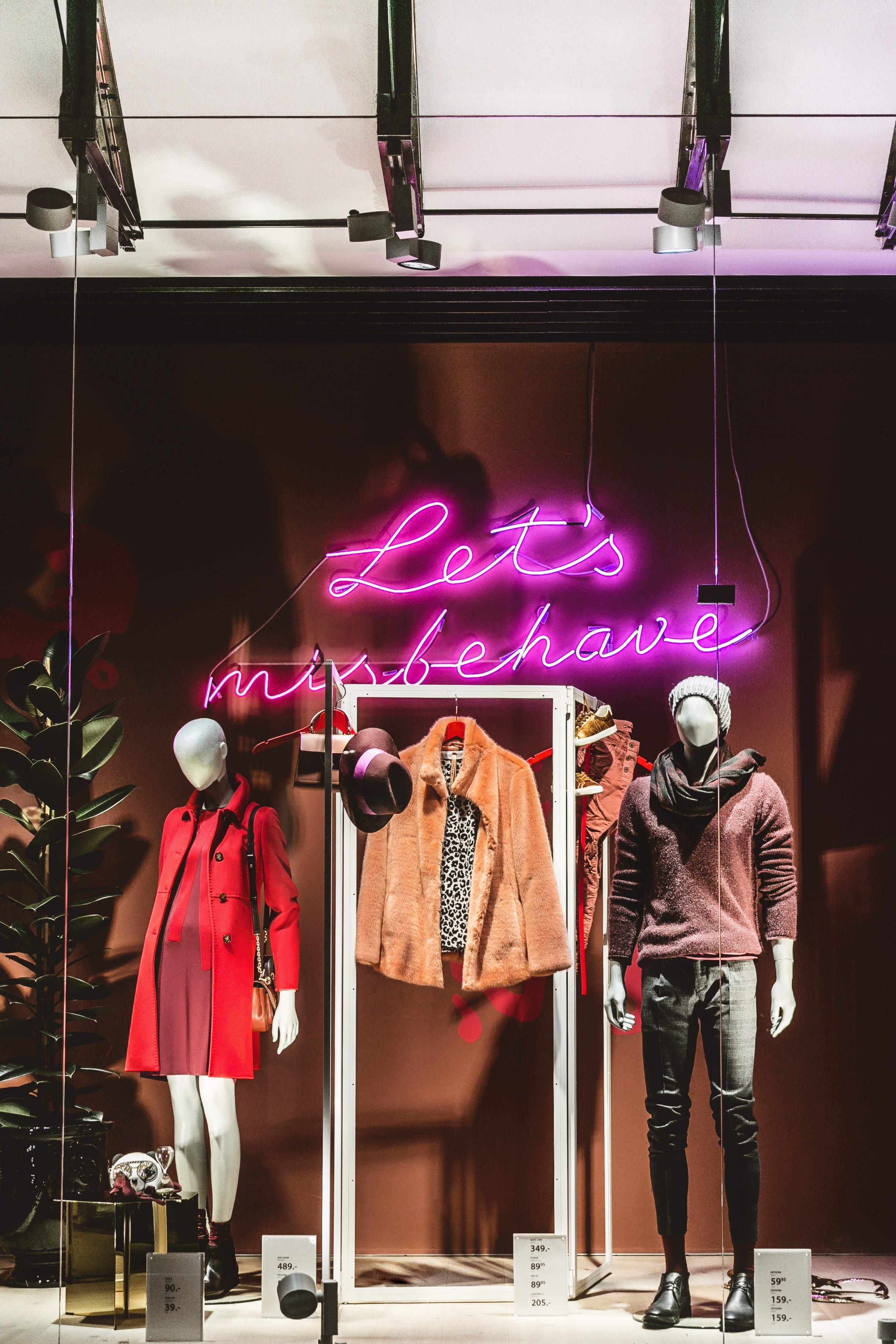 In einem Schaufenster sind unter einem Neonschild mit der Aufschrift „Lasst uns uns daneben benehmen“ Schaufensterpuppen-Outfits zu sehen. Die linke Schaufensterpuppe trägt einen roten Mantel und Stiefel, während die rechte Schaufensterpuppe eine Pelzjacke, Jeans und einen Schal trägt. Auf der Kleiderstange thront ein Hut.