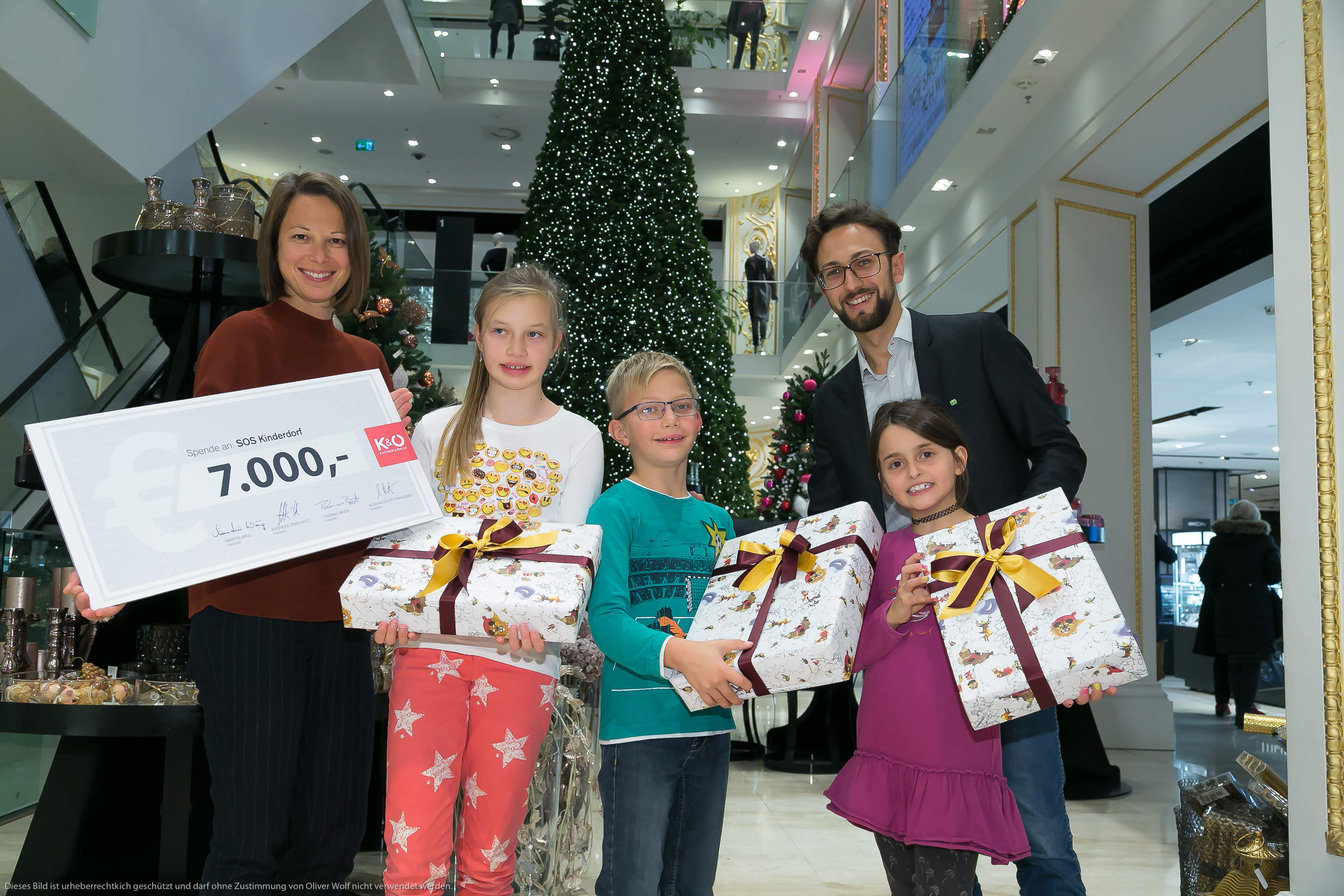Eine Frau und ein Mann stehen in einem Geschäft und halten einen riesigen Scheck über 7.000 Euro in der Hand. Drei lächelnde Kinder halten eingepackte Geschenke in der Hand. Im Hintergrund ist ein großer Weihnachtsbaum mit Ornamenten und festlicher Dekoration zu sehen.