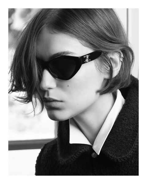 Schwarz-Weiß-Foto einer Person mit kurzem Haar, die eine dunkle Sonnenbrille und ein Hemd mit Kragen unter einer strukturierten Jacke trägt und in einer schwach beleuchteten Umgebung zur Seite schaut.