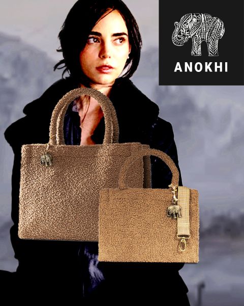 Im Hintergrund steht eine Frau mit kurzen dunklen Haaren. Im Vordergrund sind zwei strukturierte braune Handtaschen mit Elefantenanhängern zu sehen, und in der rechten oberen Ecke erscheint ein "ANOKHI"-Logo mit einem stilisierten Elefanten.