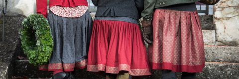 Drei festlich gekleidete Personen stehen nebeneinander und halten einen grünen Kranz in den Händen. Sie tragen gemusterte Röcke in Rot- und Dunkelgrautönen, warme Jacken und Handschuhe. Die Szene scheint sich im Freien auf Steinstufen abzuspielen.