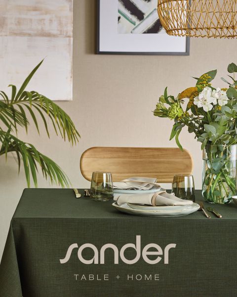 Ein moderner Esstisch mit einer grünen Tischdecke, Tellern, Gläsern, Servietten, einem Blumenarrangement und der Aufschrift „sander TABLE + HOME“.