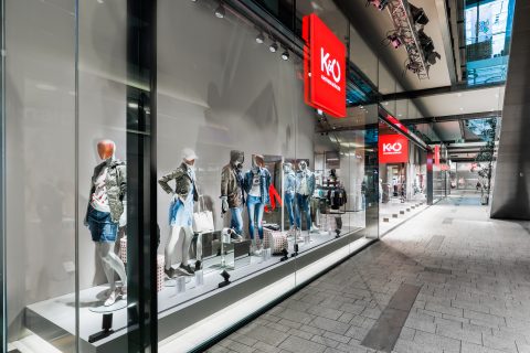Schaufenster mit Schaufensterpuppen in Freizeitkleidung wie Jacken und Shorts. Das Geschäft hat große Glasfenster mit einem roten „KO“-Schild darüber. Das moderne Interieur zeichnet sich durch helle Beleuchtung und verschiedene ausgestellte Accessoires aus.