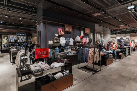 Ein modernes Interieur eines Bekleidungsgeschäfts mit ordentlich angeordneten Regalen und Ablagen, auf denen verschiedene Kleidungsstücke ausgestellt sind. Das Geschäft bietet verschiedene Marken, eine Vielzahl von Kleidungsstilen und Accessoires bei heller, stimmungsvoller Beleuchtung. Der Boden ist gefliest und die Decke ist im Industriestil gehalten.