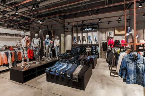 In einem modernen Bekleidungsgeschäft tragen Schaufensterpuppen legere Outfits. Verschiedene Jeans und Jacken sind ordentlich auf Regalen und Ständern ausgestellt. Das Geschäft ist gut beleuchtet und verfügt über Abteilungen für verschiedene Bekleidungsstile, darunter einen „Slim Fit“-Bereich.