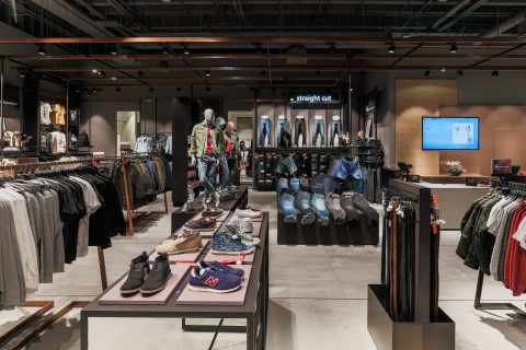 Ein modernes Bekleidungsgeschäft stellt auf Ständern und Tischen verschiedene Kleidungsstücke aus, darunter Jeans, Hemden und Jacken. Schaufensterpuppen präsentieren Outfits, und Schuhe stehen auf einem zentralen Tisch. Das Geschäft wirkt elegant und übersichtlich, an der Wand hängt ein Fernsehbildschirm.
