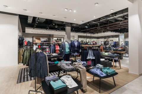 Ein modernes Interieur eines Bekleidungsgeschäfts mit ordentlich angeordneten Regalen und Tischen, auf denen verschiedene Kleidungsstücke wie Hemden, Jacken und Hosen ausgestellt sind. Der Raum zeichnet sich durch helle Beleuchtung und eine neutrale Farbpalette mit Holzakzenten aus.