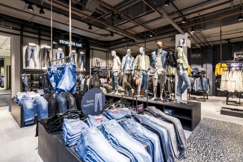 Ein Schaufenster in einem Bekleidungsgeschäft mit Stapeln gefalteter Jeans in verschiedenen Stilen und Farben. Im Hintergrund sind Schaufensterpuppen in lässiger Kleidung, darunter Jeansjacken und -hosen, angeordnet. Helles, gut beleuchtetes Interieur mit modernem Design.