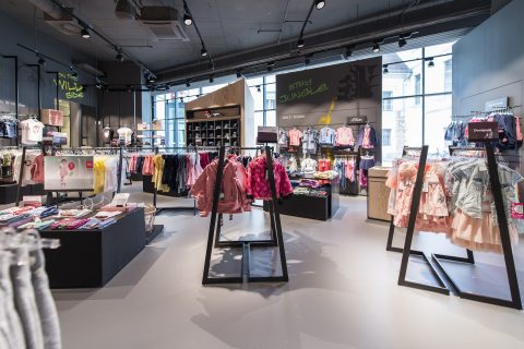 Ein hell erleuchtetes Kinderbekleidungsgeschäft, das eine Vielzahl farbenfroher Kleidungsstücke präsentiert. Im Inneren befinden sich Kleiderständer mit Kleidern, Jacken und Hemden. Große Fenster lassen natürliches Licht in den modernen Raum fallen.