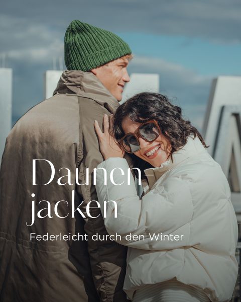 Eine lächelnde Frau in einer weißen Pufferjacke und Sonnenbrille lehnt sich an einen Mann mit grüner Mütze und braunem Mantel. Sie befinden sich im Freien mit großen Metallbuchstaben im Hintergrund. Der Text lautet: "Daunenjacken - Federleicht durch den Winter.