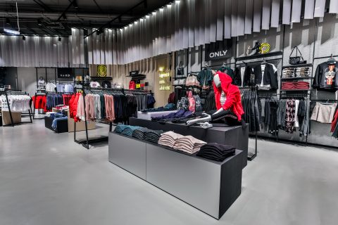 Ein modernes Bekleidungsgeschäft mit ordentlich angeordneten Ständern voller farbenfroher Kleidungsstücke, darunter Pullover, Jacken und Hosen. Das Geschäft zeichnet sich durch elegante Auslagen und helle Beleuchtung aus, die die farbenfrohen Kleidungsstücke, die zum Verkauf angeboten werden, hervorheben.