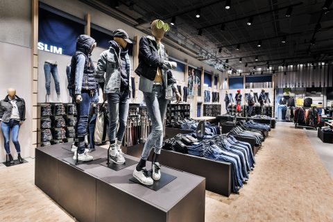 Ein modernes Interieur eines Bekleidungsgeschäfts mit Schaufensterpuppen in Jeans und Jacken. Auf Regalen und Ständern werden gefaltete und hängende Jeans ausgestellt, mit dem Hinweis „SLIM“. Das Geschäft hat ein elegantes, industrielles Design mit Holzboden.