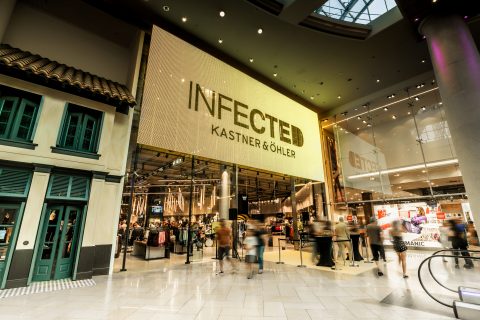 Ein belebter Einkaufsbereich im Innenbereich mit einem großen, beleuchteten Schild mit der Aufschrift „INFECTED KASTNER & ÖHLER“. Die Käufer bewegen sich durch den Raum und sehen verschiedene Geschäfte, darunter eines mit traditioneller Fassade auf der linken Seite und ein anderes mit modernem Design.