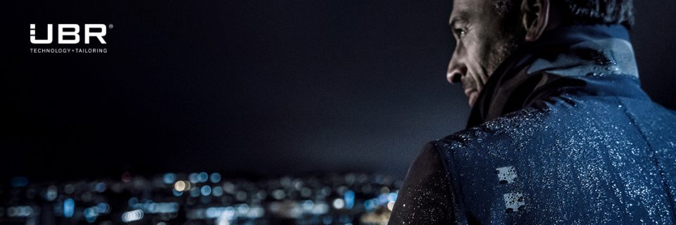 Eine Person in einer Jacke blickt über die Schulter auf eine nächtliche Stadtlandschaft. Auf der Jacke sind Wassertropfen, die die Lichter der Stadt reflektieren. In der oberen linken Ecke sind das UBR-Logo und die Worte „Technology Tailoring“ zu sehen.