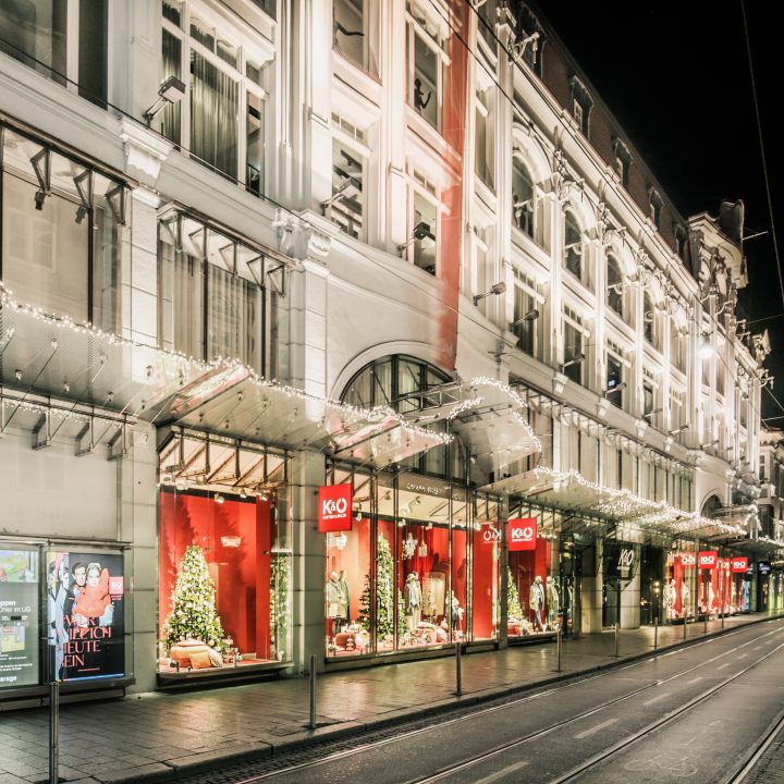 Ein hell erleuchtetes Kaufhaus bei Nacht, geschmückt mit Weihnachtsdekorationen. Das Gebäude verfügt über große Fenster, die festliche Szenen mit roten Akzenten und Weihnachtsbäumen zeigen. Die Straße ist leer und die Fassade leuchtet mit dekorativen Lichtern.