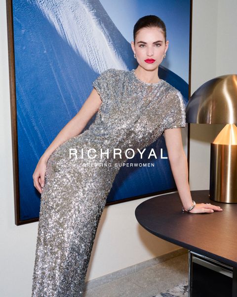 Eine Frau in einem funkelnden silbernen Kleid posiert neben einem Schreibtisch und einer modernen Lampe, mit einem großen abstrakten blauen und weißen Kunstwerk im Hintergrund. Der Text auf dem Bild lautet "RICH&ROYAL DRESSING SUPERWOMEN.".