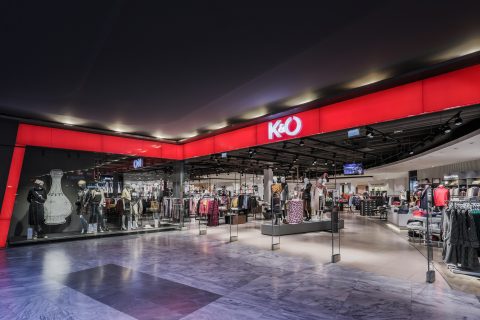 Das Bild zeigt das Äußere eines K&O-Bekleidungsgeschäfts in einem Einkaufszentrum. Die Ladenfront ist in Rot und Schwarz gehalten und mit Schaufensterpuppen versehen, die verschiedene Outfits präsentieren. Im Inneren sind Kleiderständer neben heller Deckenbeleuchtung zu sehen.