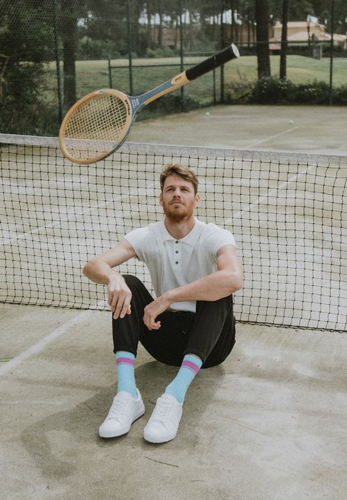 Ein Mann mit roten Haaren und Bart sitzt lässig vor einem Tennisnetz auf einem Platz. Er trägt ein weißes Hemd, eine schwarze Hose, bunte Socken und weiße Turnschuhe. Über ihm fliegt ein hölzerner Tennisschläger durch die Luft.