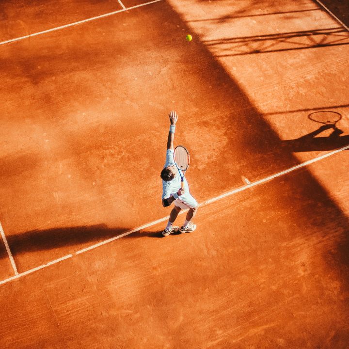 Ein Tennisspieler bereitet sich auf einem sonnenbeschienenen Sandplatz auf seinen Aufschlag vor. Der Schatten des Spielers und seines Schlägers ist auf der Oberfläche des Platzes sichtbar und unterstreicht die sportliche Bewegung. Die Szene fängt die dynamische Energie des Sports ein.