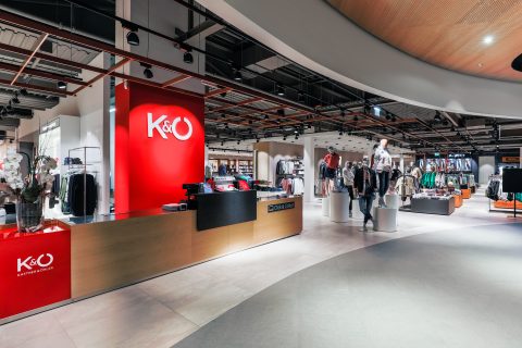 Inneneinrichtung eines Bekleidungsgeschäfts mit modernem Design. Eine leuchtend rote Wand zeigt das Logo des Geschäfts und eine Kasse ist sichtbar. Schaufensterpuppen präsentieren Kleidung und Kleiderständer voller Kleidung füllen den Hintergrund.