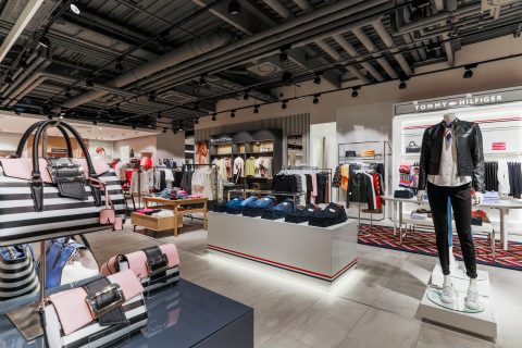 Ein modisches Bekleidungsgeschäft mit moderner Beleuchtung. In den Auslagen sind Hemden, Jacken und Accessoires, darunter gestreifte Handtaschen, zu sehen. Schaufensterpuppen sind in trendige Outfits gekleidet. An der Wand ist die Marke Tommy Hilfiger zu sehen.