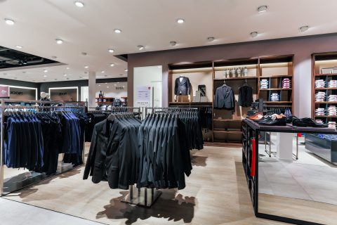 Ein modernes Interieur eines Bekleidungsgeschäfts mit ordentlich angeordneten Kleiderständern voller schwarzer Anzüge und einer Rückwand mit Regalen, in denen gefaltete Hemden und Anzüge ausgestellt sind. Der Raum ist gut beleuchtet und wird durch Scheinwerfer hervorgehoben, die die Präsentation hervorheben.