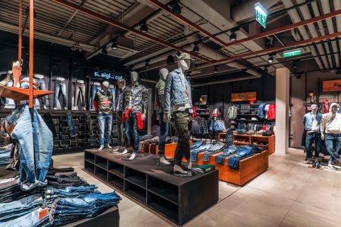 Ein modernes Interieur eines Bekleidungsgeschäfts mit Schaufensterpuppen in lässigen Jeans-Outfits. Auf Regalen und Ständern werden gefaltete Jeans und Jacken ausgestellt. Der Raum ist gedämpft beleuchtet und im Industriestil mit sichtbaren Deckenrohren eingerichtet. Im Hintergrund stöbern Käufer.