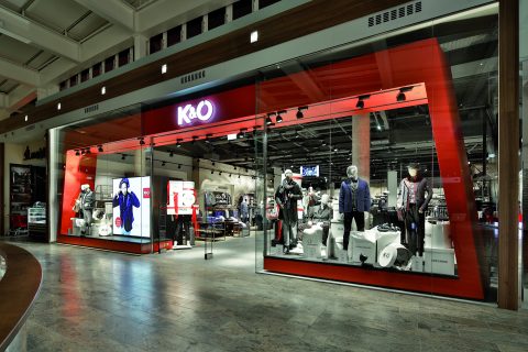 Schaufenster eines Bekleidungsgeschäfts mit Schaufensterpuppen, die verschiedene Outfits in einem hell erleuchteten Einkaufszentrum präsentieren. Der Eingang ist breit, in den Farben Rot und Schwarz gehalten und das Logo des Geschäfts ist gut sichtbar über der Tür angebracht.