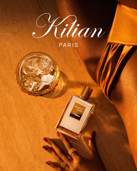 Ein Glas Whisky mit Eis, ein Parfümflakon mit der Aufschrift „Kilian Paris“ und eine Frauenhand mit goldenen Ringen liegen auf einer Marmorplatte neben einer Frau in goldenem Satinkleid. Warmes, goldenes Licht sorgt für eine luxuriöse Stimmung.