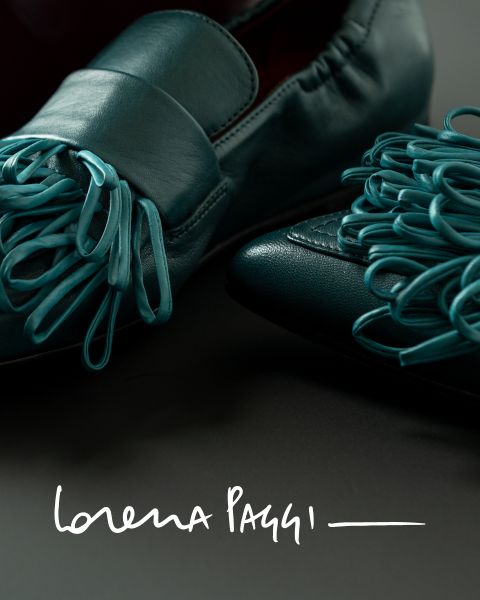Nahaufnahme von blaugrünen Lederschuhen mit dekorativen Schlaufen oben in Zehennähe, auf einer dunklen Oberfläche platziert. Das Bild zeigt unten den Text „Lorena Paggi“ in weißer Schrift.