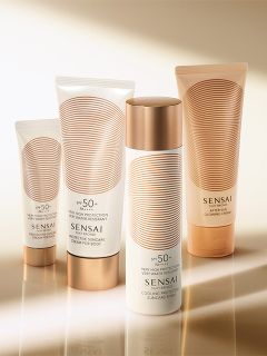 Vier Sensai-Sonnenpflegeprodukte mit weißer und goldener Verpackung werden auf einer reflektierenden Oberfläche mit einem warmen, leuchtenden Hintergrund präsentiert.