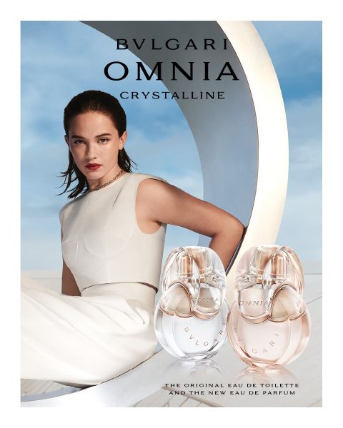 Eine Frau in einem weißen Kleid lehnt sich vor einer modernen Architekturkulisse unter blauem Himmel zurück. Im Vordergrund stehen zwei Flakons des Parfums Bvlgari Omnia Crystalline, die klare und bernsteinfarbene Farbtöne aufweisen.
