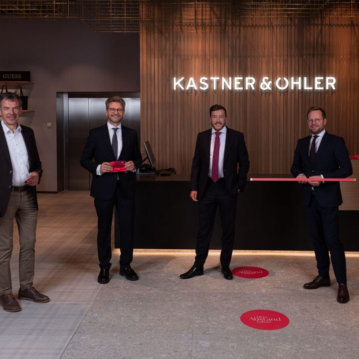 Vier Männer in formeller Kleidung stehen in einem modernen Ladenlokal vor einem Tresen mit einem „Kastner & Öhler“-Schild. Zwei halten ein Band in der Hand, möglicherweise für eine feierliche Eröffnung. Das Interieur ist von schlichten, minimalistischen Designelementen geprägt.