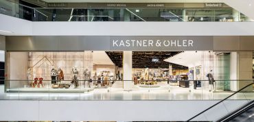 Weitwinkelansicht des Interieurs eines modernen Kastner & Öhler-Geschäfts. Die Schaufensterfront mit ihren großen Glasfenstern zeigt Schaufensterpuppen, die Modekleidung präsentieren. Der Name des Geschäfts ist oben gut sichtbar angebracht. Im Obergeschoss sind weitere Markenschilder angebracht.