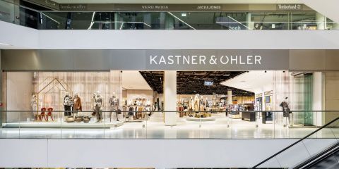 Weitwinkelansicht des Interieurs eines modernen Kastner & Öhler-Geschäfts. Die Schaufensterfront mit ihren großen Glasfenstern zeigt Schaufensterpuppen, die Modekleidung präsentieren. Der Name des Geschäfts ist oben gut sichtbar angebracht. Im Obergeschoss sind weitere Markenschilder angebracht.