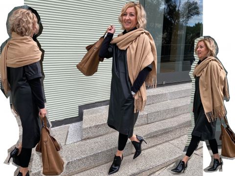 Eine Frau mit lockigem blondem Haar trägt ein schwarzes Kleid, schwarze Leggings und einen hellbraunen Schal. Sie trägt eine braune Handtasche und geht lächelnd und nach vorne blickend die Stufen vor einer gerippten Gebäudefassade hinauf.