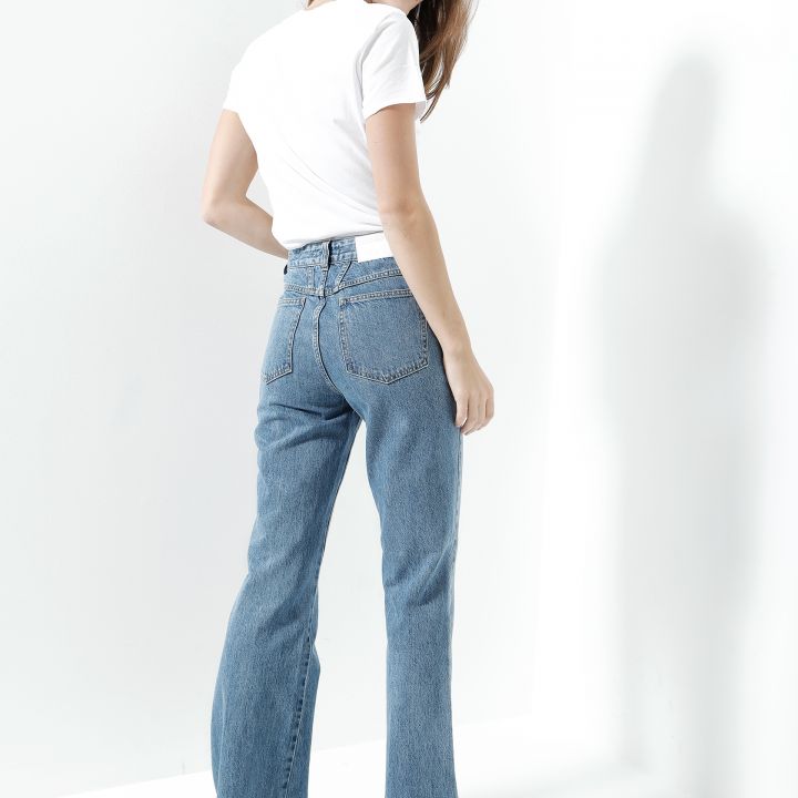 Eine Frau in weißem T-Shirt und Blue Jeans steht vor einem weißen Hintergrund und dreht sich leicht, um über ihre Schulter zu blicken. Sie trägt weiße Schuhe und hat langes braunes Haar. Die Jeans hat eine hohe Taille und ein ausgestelltes Design.