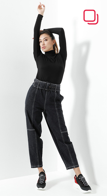 Eine Person in einem schwarzen Langarmshirt und dunklen Jeans mit hoher Taille posiert mit einem erhobenen Arm vor einem weißen Hintergrund. Sie trägt schwarze und rote Turnschuhe. Der Schatten ihrer Pose ist an der Wand sichtbar.