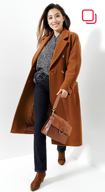 Eine Person in einem langen braunen Mantel, einer gemusterten Bluse und dunklen Jeans hält eine braune Handtasche und geht selbstbewusst. Sie hat lange Haare und trägt braune Stiefel. Der Hintergrund ist schlicht weiß.